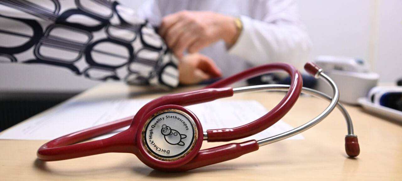 Ein Arzt misst in einer Bereitschaftspraxis in einem Krankenhaus in einem Untersuchungsraum bei einer Patientin den Blutdruck (gestellte Szene). Im Vordergrund liegt ein Stethoskop. (zu dpa: «Bürgermeister laufen Sturm gegen Pläne zu Notfallpraxen»)