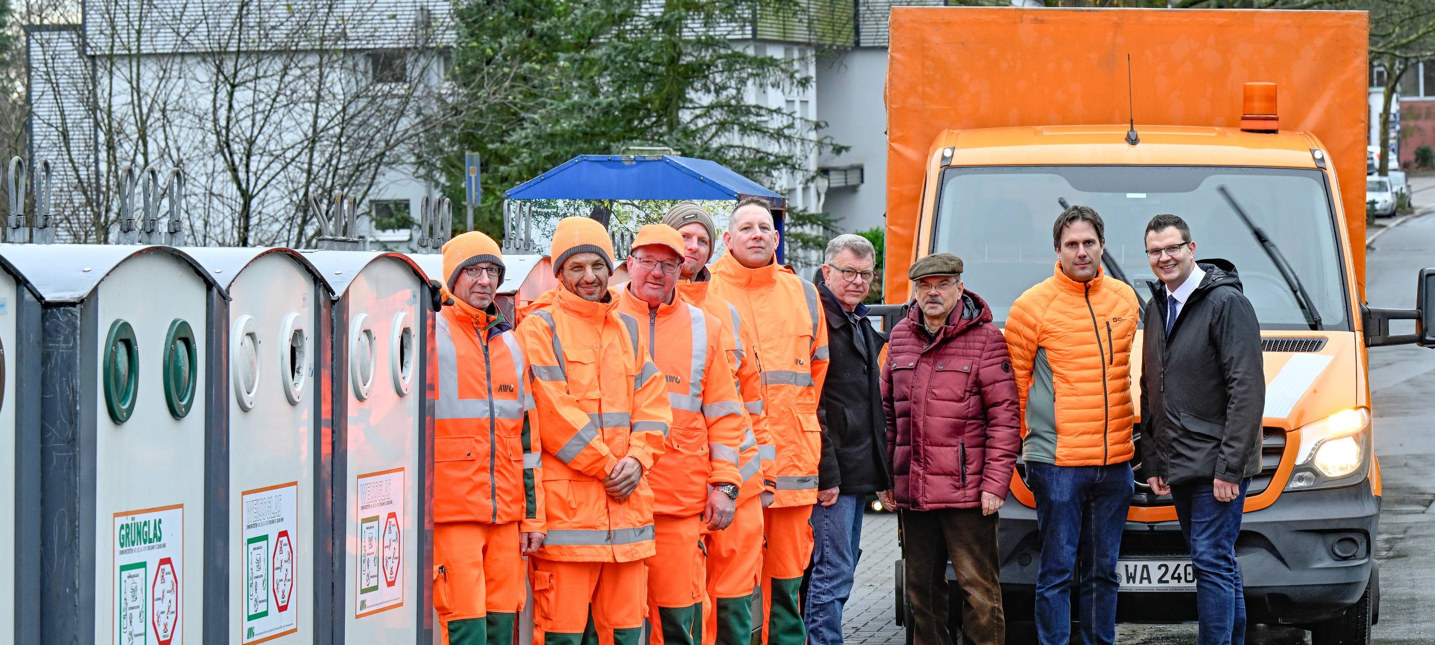 Mehrere Männer in orangefarbenen Anzügen stehen neben Müllcontainern, außerdem die Chefs der AWG
