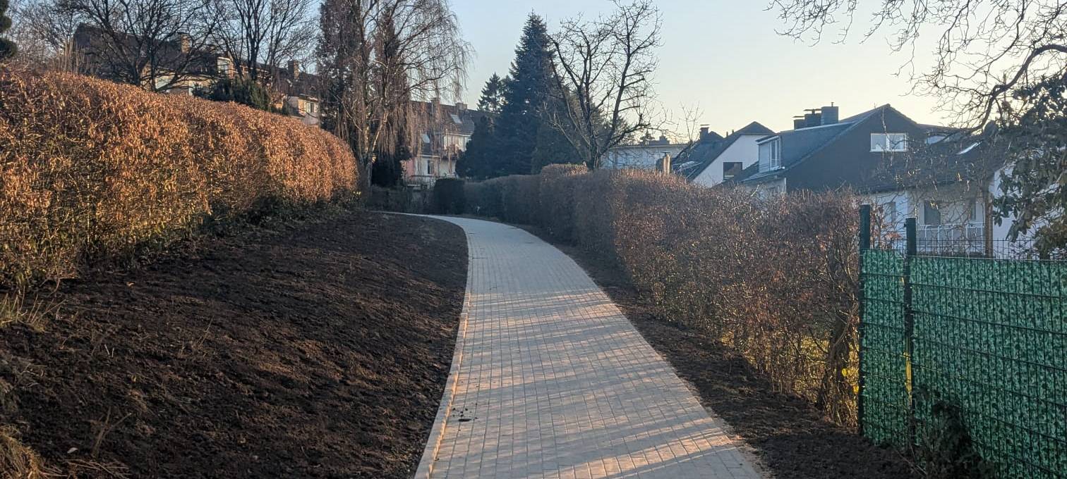 Ein gepflasterter Weg zwischen Hecken und Zäunen, im Hintergrund stehen Einfamilienhäuser